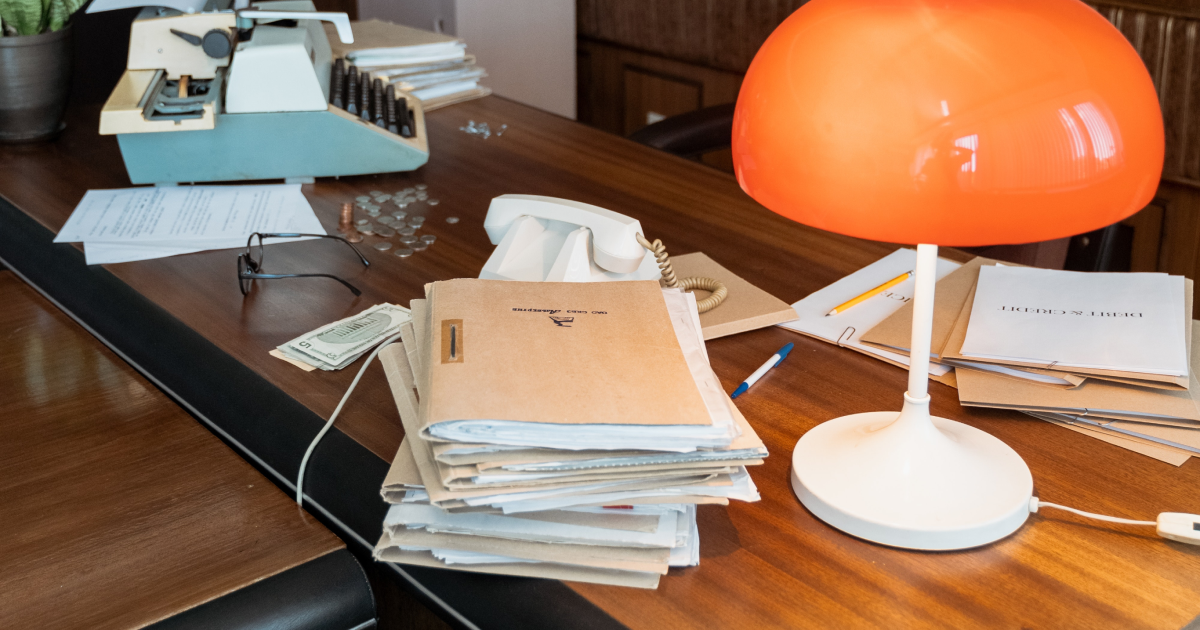 Pile of stuffed folders on desk with lamp and old typewriter in background.