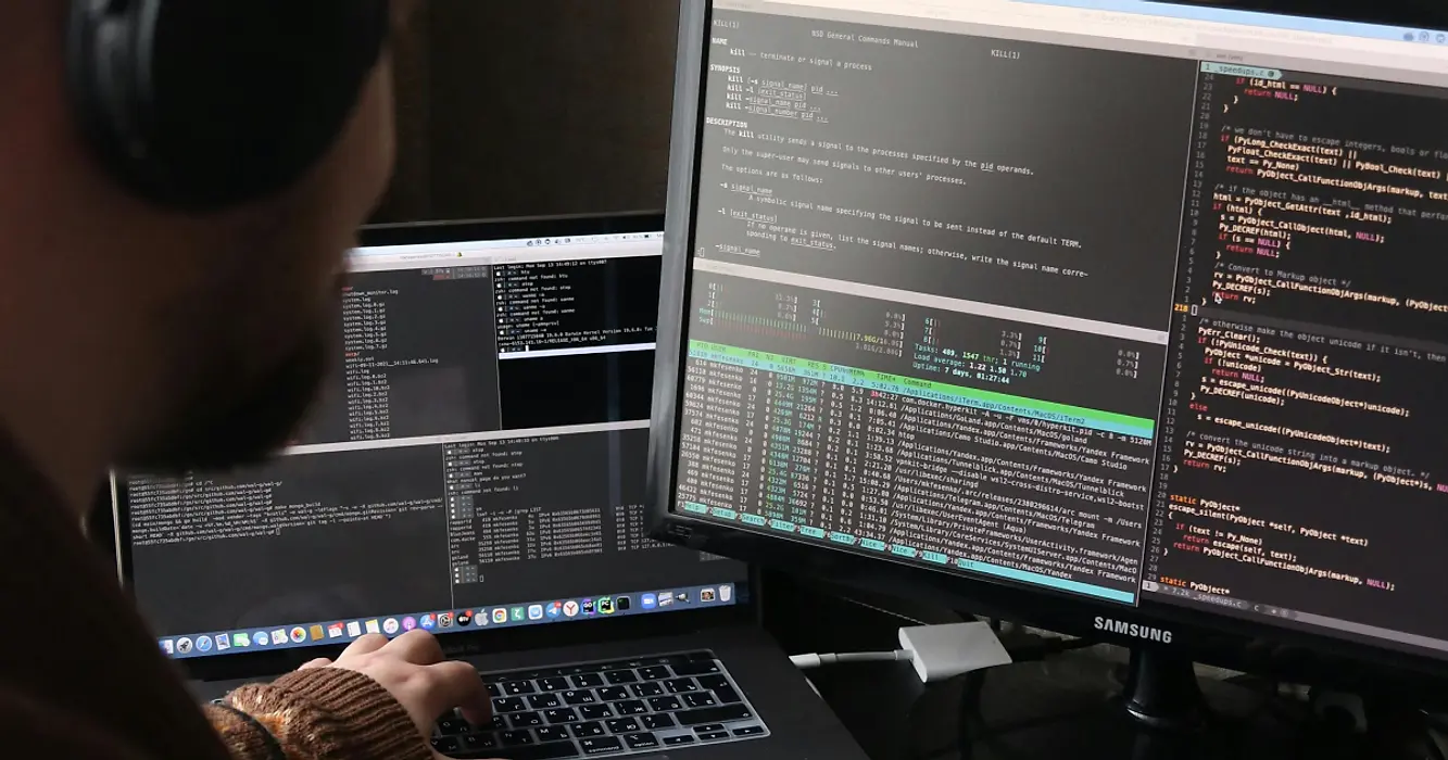 Over-shoulder view of software engineer wearing headphones and working across two monitors displaying code.