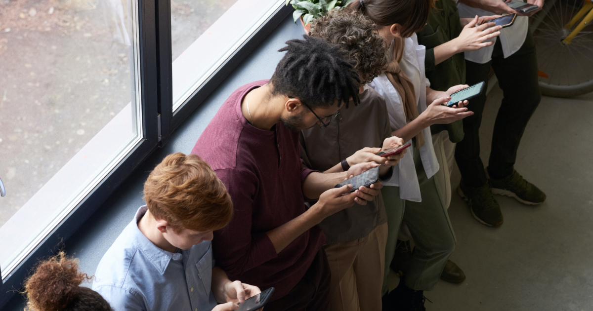 Line of people standing by window looking down at their smartphones and typing.