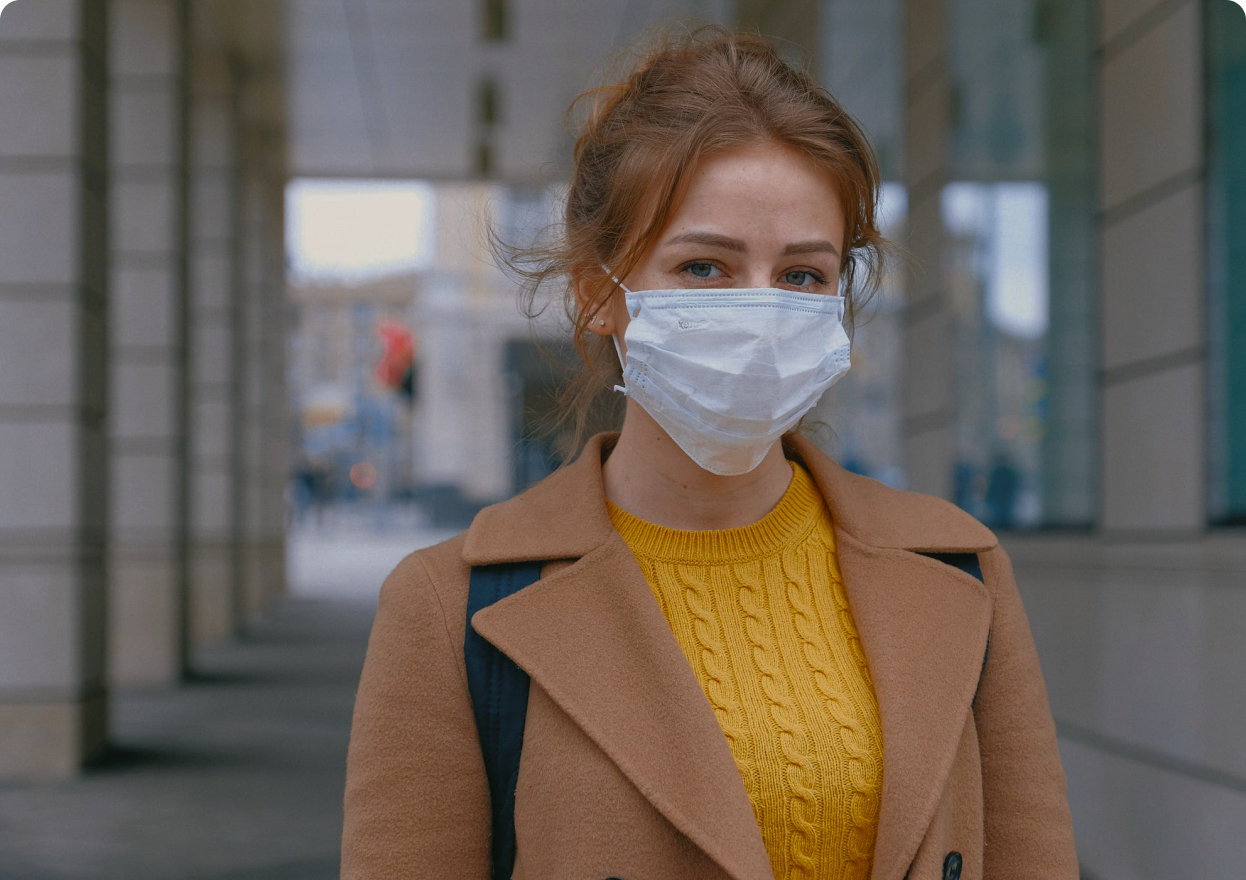 Young woman standing outside building in yellow sweater and tan coat wearing surgical mask and looking back at viewer.