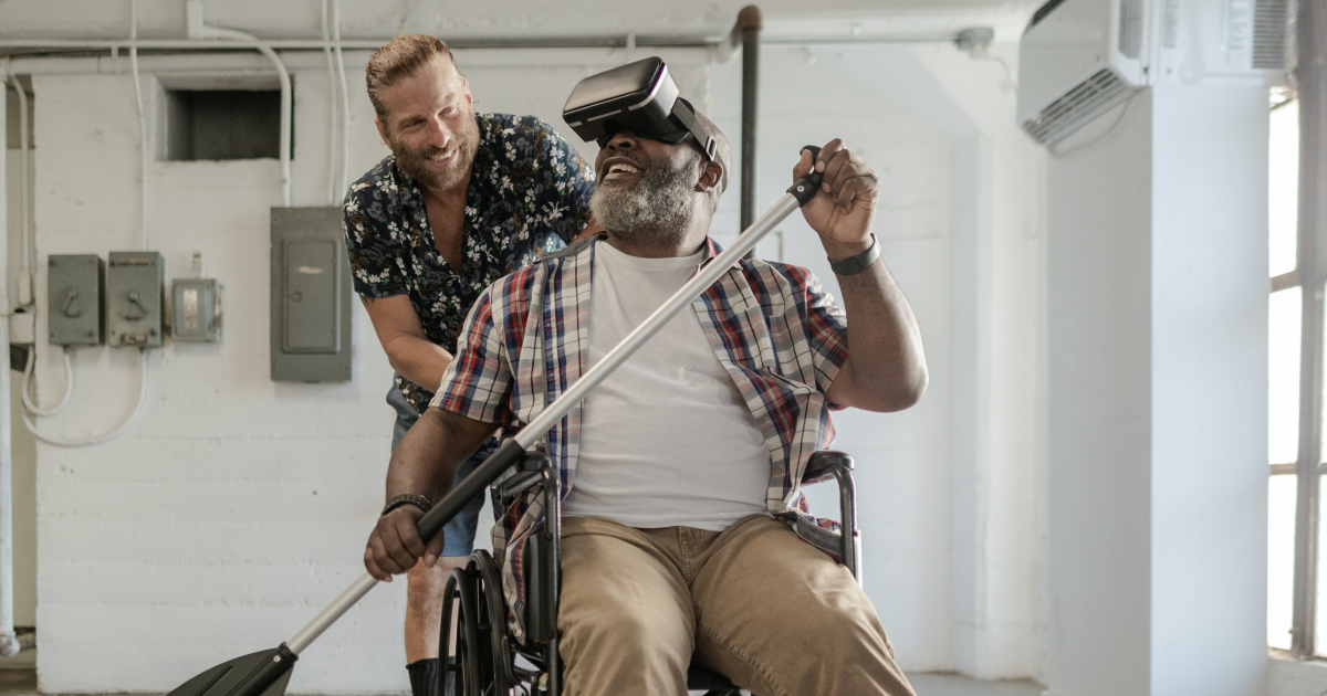 Man sitting in wheelchair wearing VR headset and rowing with canoe paddle, with smiling physical therapist behind him.