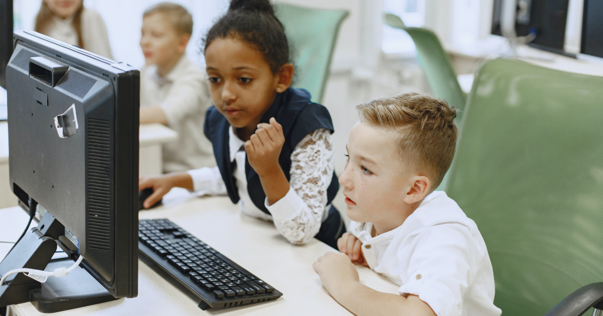 Elementary school ages girl an boy sitting in computer lab looking at the same monitor with interest.