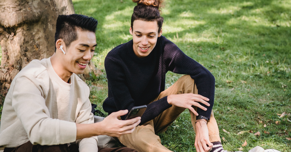 Two young men sitting outside under tree looking at smartphone and smiling.