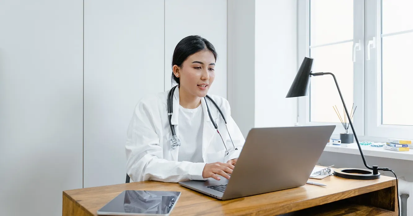 Young medical professional in white lab coat and stethoscope sitting at desk in brightly lit office working on laptop computer.