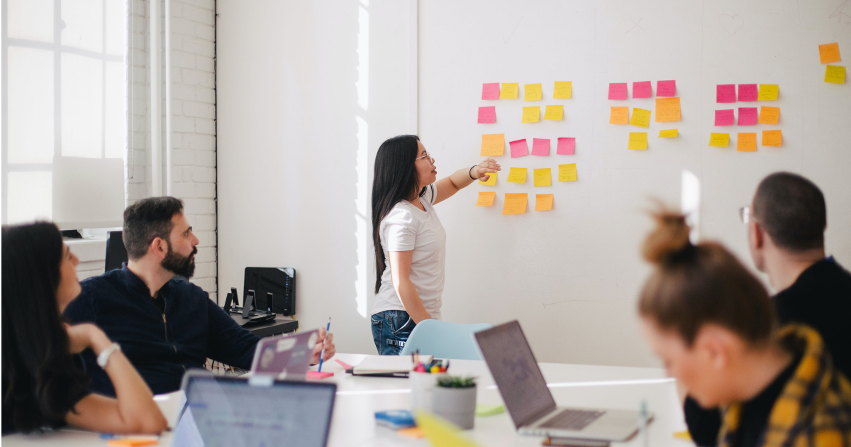 Young woman with long hair leading brainstorming session with colorful post it notes stuck to white wall.