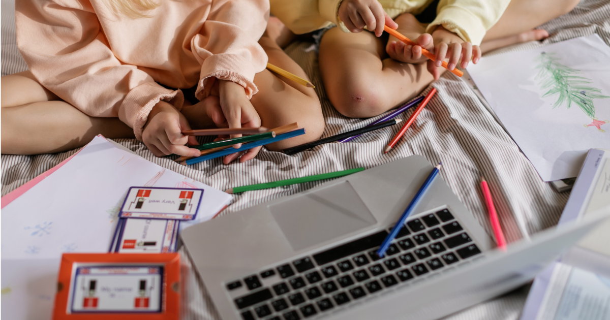 Open laptop surrounded by flashcards and art supplies with two young kids sitting looking at it.