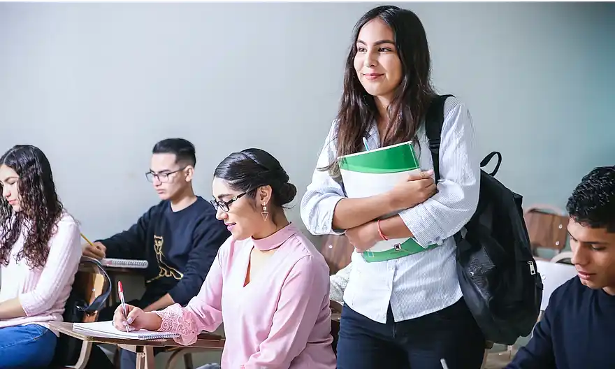 Young smiling teen with notebooks and backpack standing in classroom surrounded by other students sitting at desks taking notes.