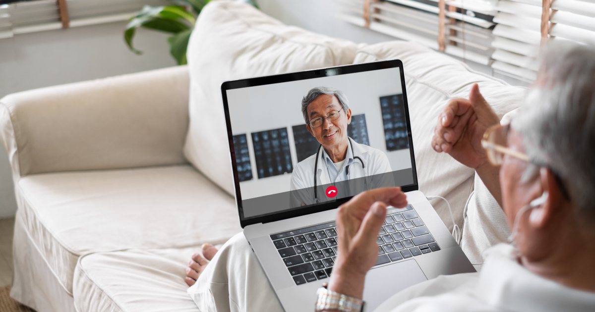 Gray-haired man siting on couch with open laptop in lap having a telehealth visit with his doctor via video call.