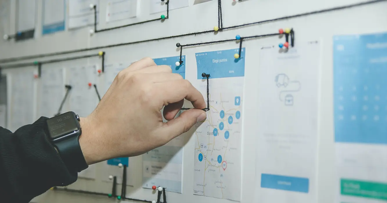 Hand using string and pins to map user journey on bulletin board covered in app screen printouts.