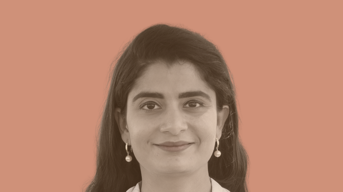 A person with long dark hair in a doctor's coat with earrings and a polka-dot shirt
