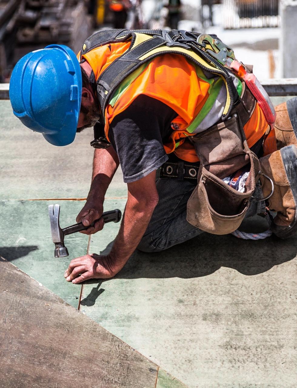 A builder in a blue hard hat and hi-vis vest hammering on a construction site