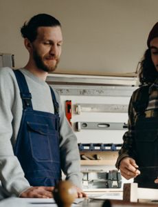 two men are standing next to each other in a workshop looking at a piece of wood .