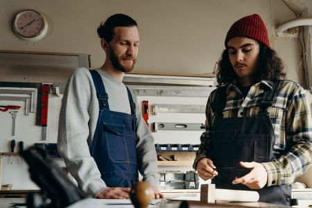 two men are standing next to each other in a workshop looking at a piece of wood .