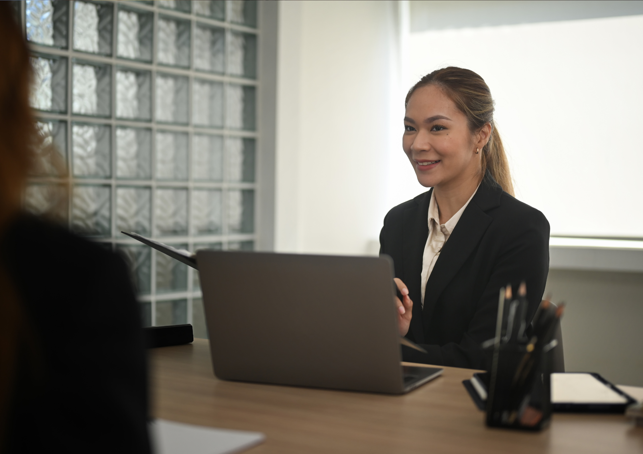 Smiling businesswoman in a suit at a desk with a laptop, in a meeting.