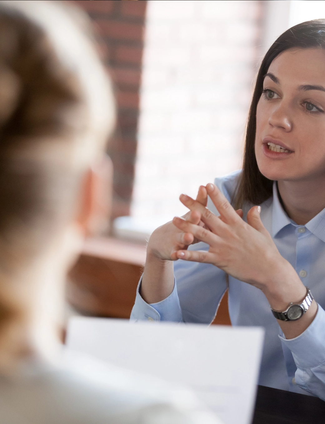 A woman in a blue shirt speaks with clasped hands across a table to another person.