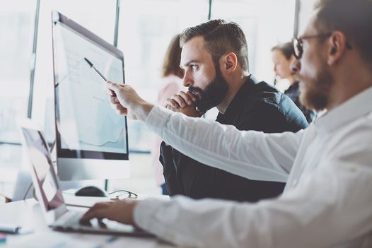 Two men review charts and data on a computer monitor in an office.