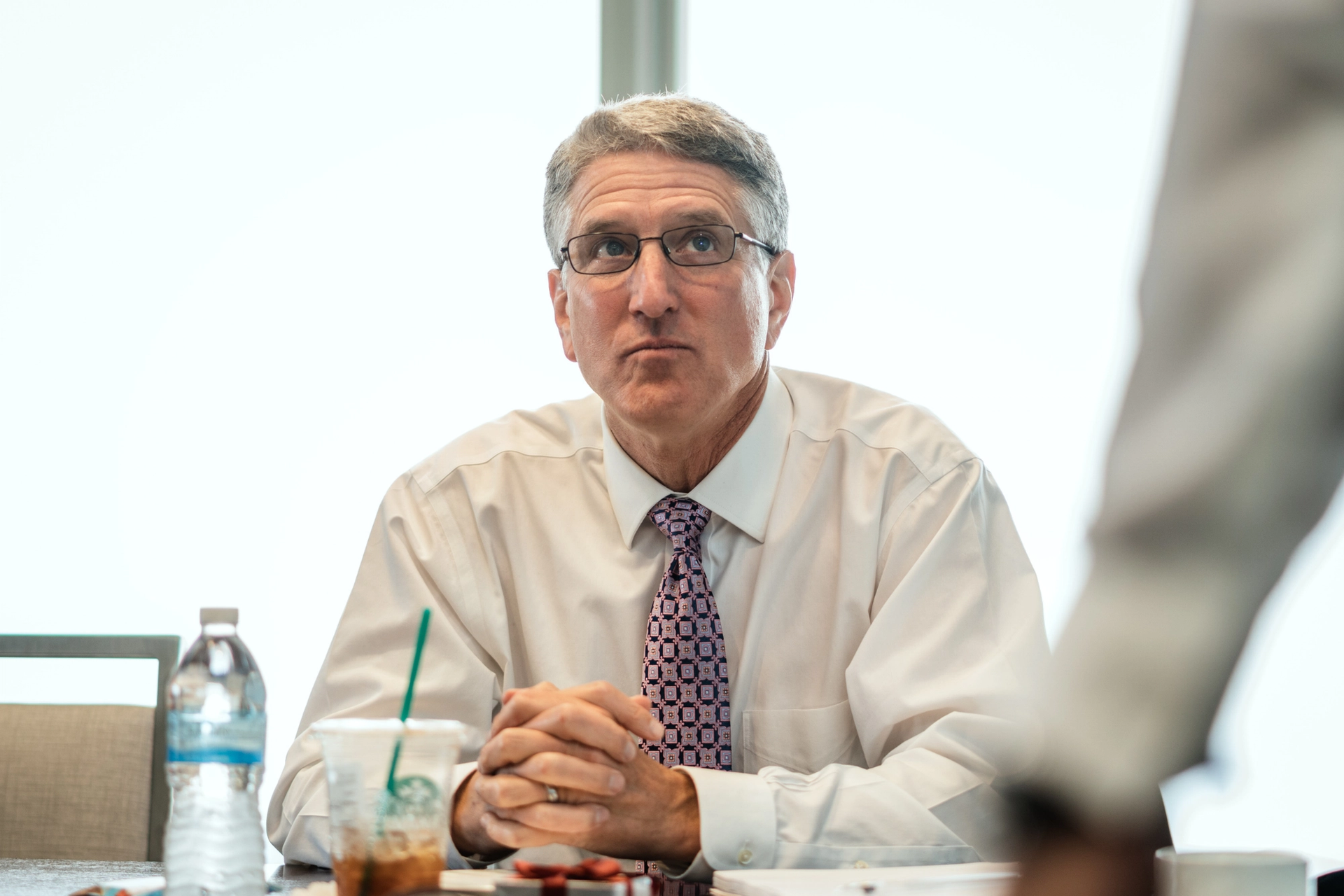 A man with gray hair and glasses in a white shirt and tie sits at a table, hands clasped, looking up. A water bottle and coffee cup are on the table.