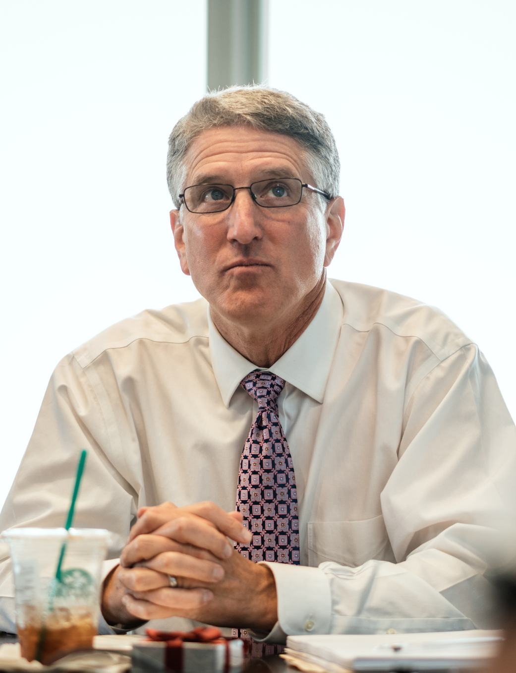 A man with gray hair and glasses in a white shirt and tie sits at a table, hands clasped, looking up. A water bottle and coffee cup are on the table.