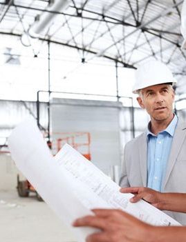 two men in hard hats are looking at a blueprint in a warehouse .