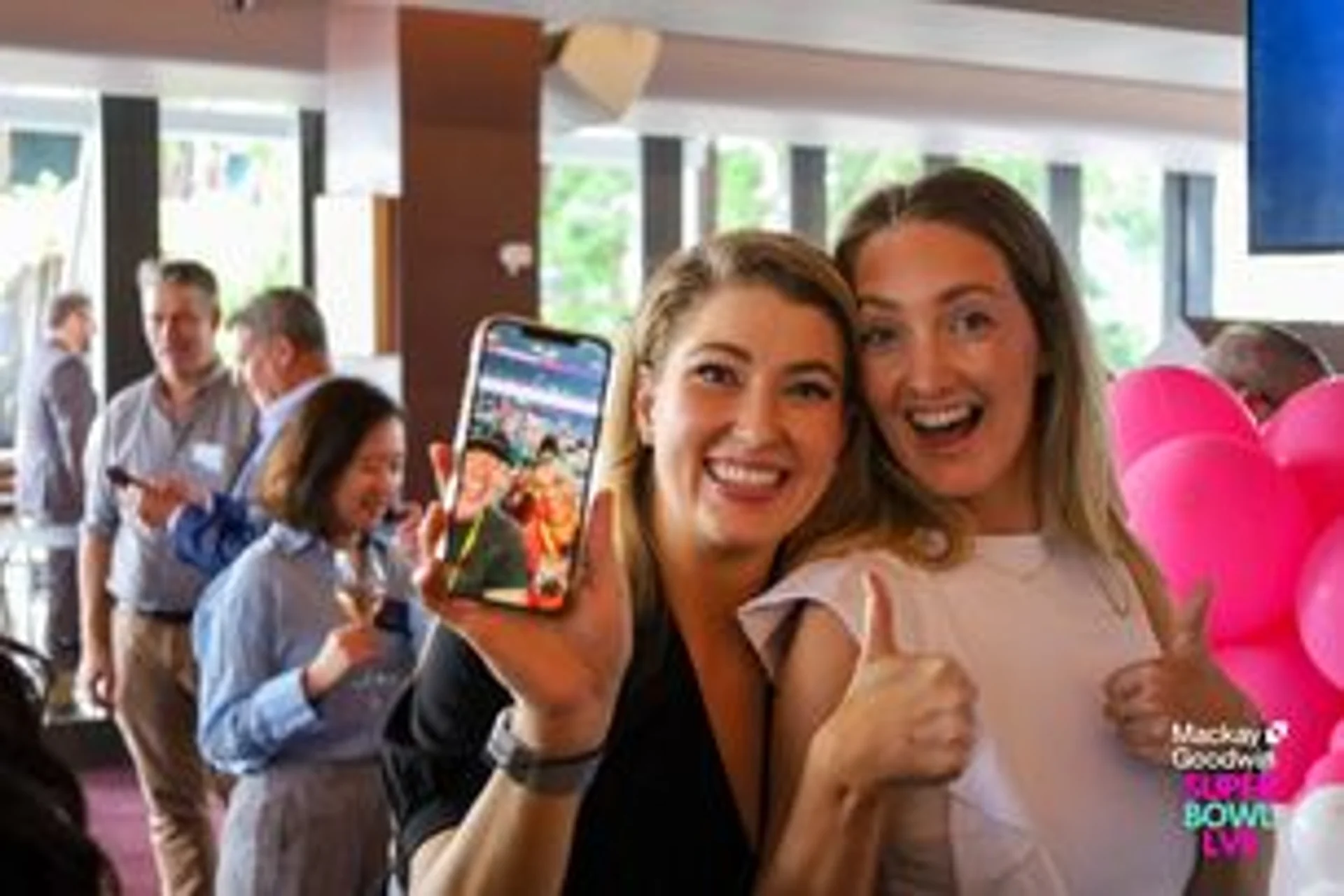 two women are posing for a picture with a cell phone and giving a thumbs up .