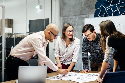 a group of people are standing around a table looking at a laptop .
