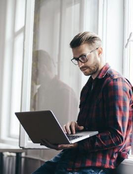 A man in glasses and a plaid shirt works on a laptop while sitting on a windowsill.