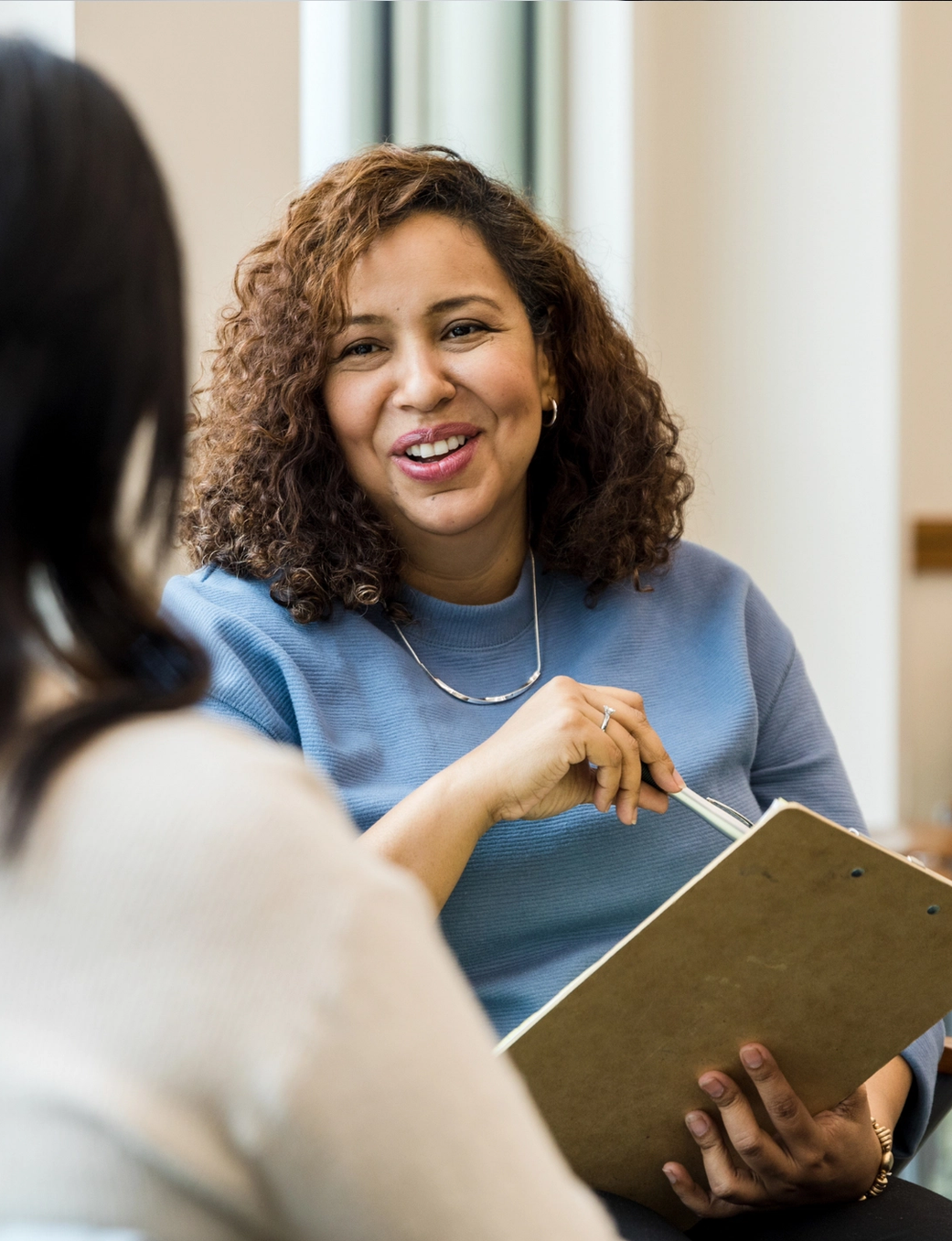 A smiling woman with curly hair holds a clipboard and pen while talking to another woman.