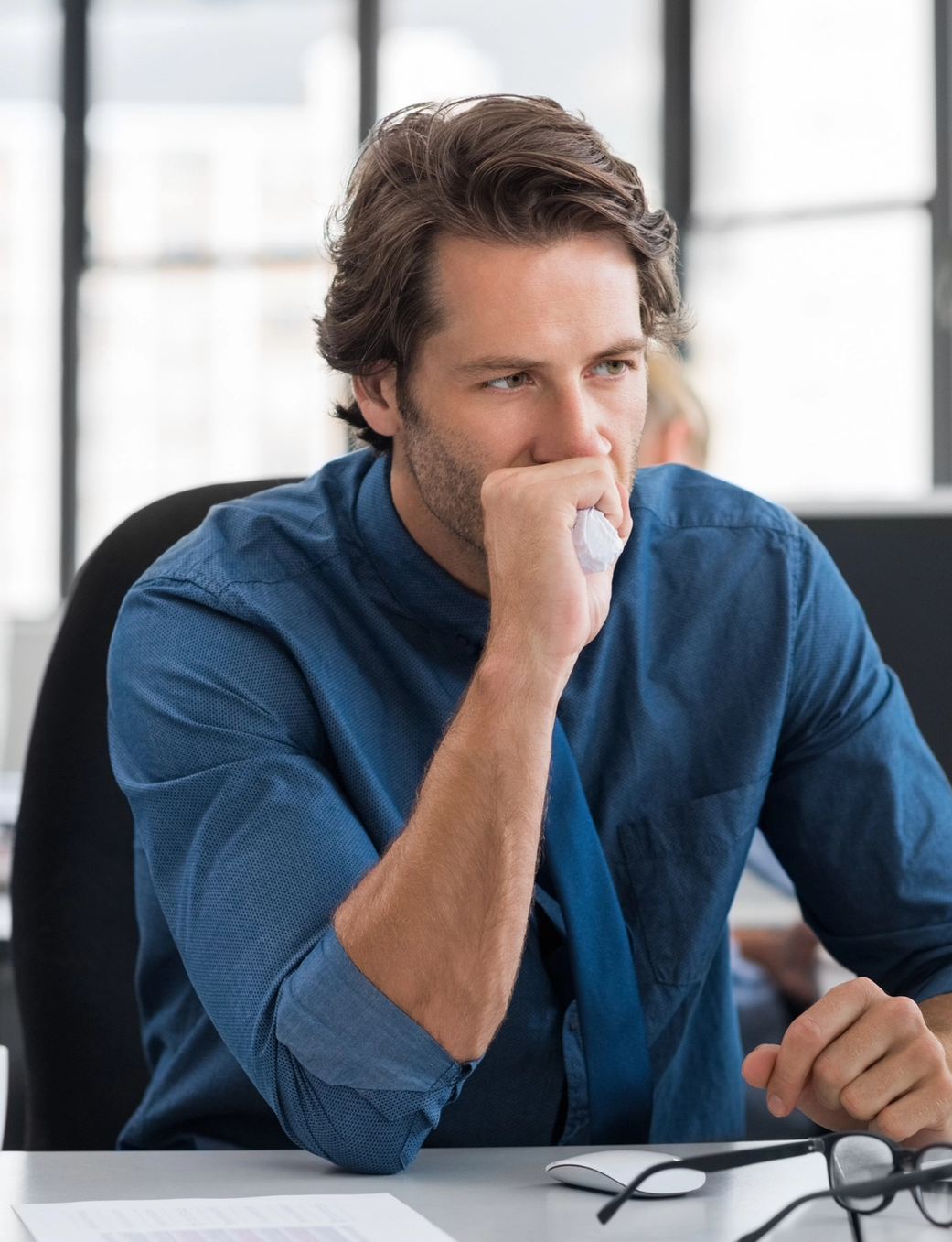A man in a blue shirt looking intently at a computer with his hand to his mouth, in an office setting.