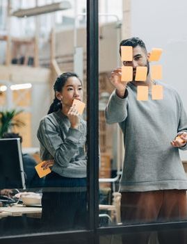 a man and a woman are looking at sticky notes on a glass wall .