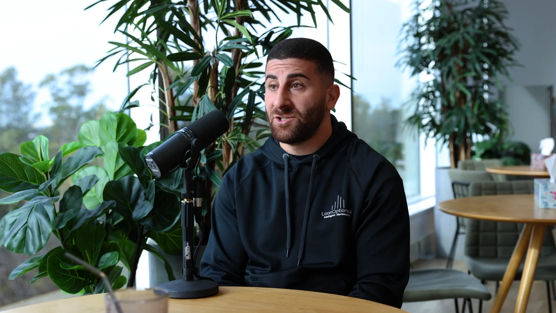 A man with a beard speaks into a microphone, wearing a black hoodie, in a bright room with windows and plants.