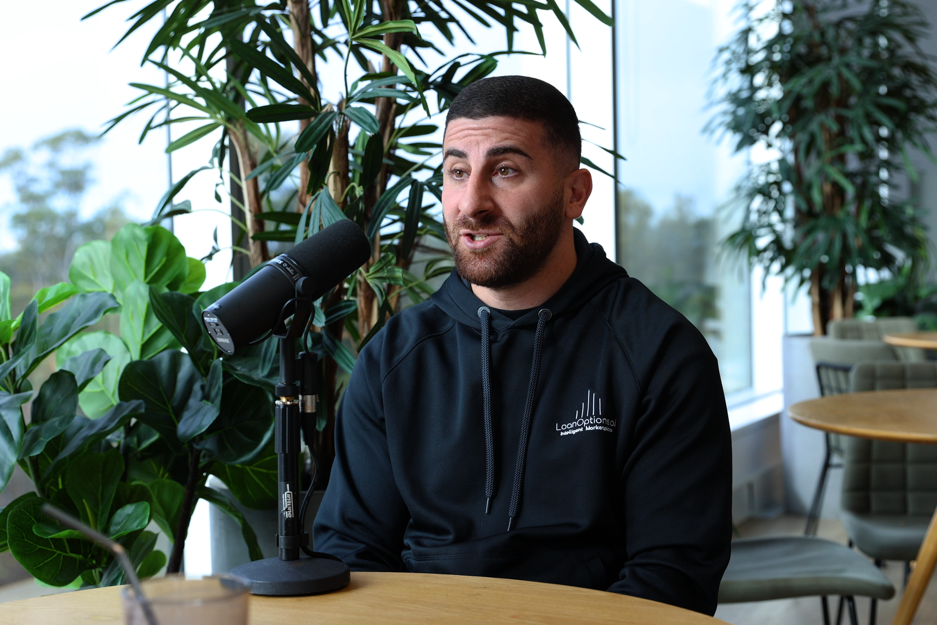 A man with a beard speaks into a microphone, wearing a black hoodie, in a bright room with windows and plants.
