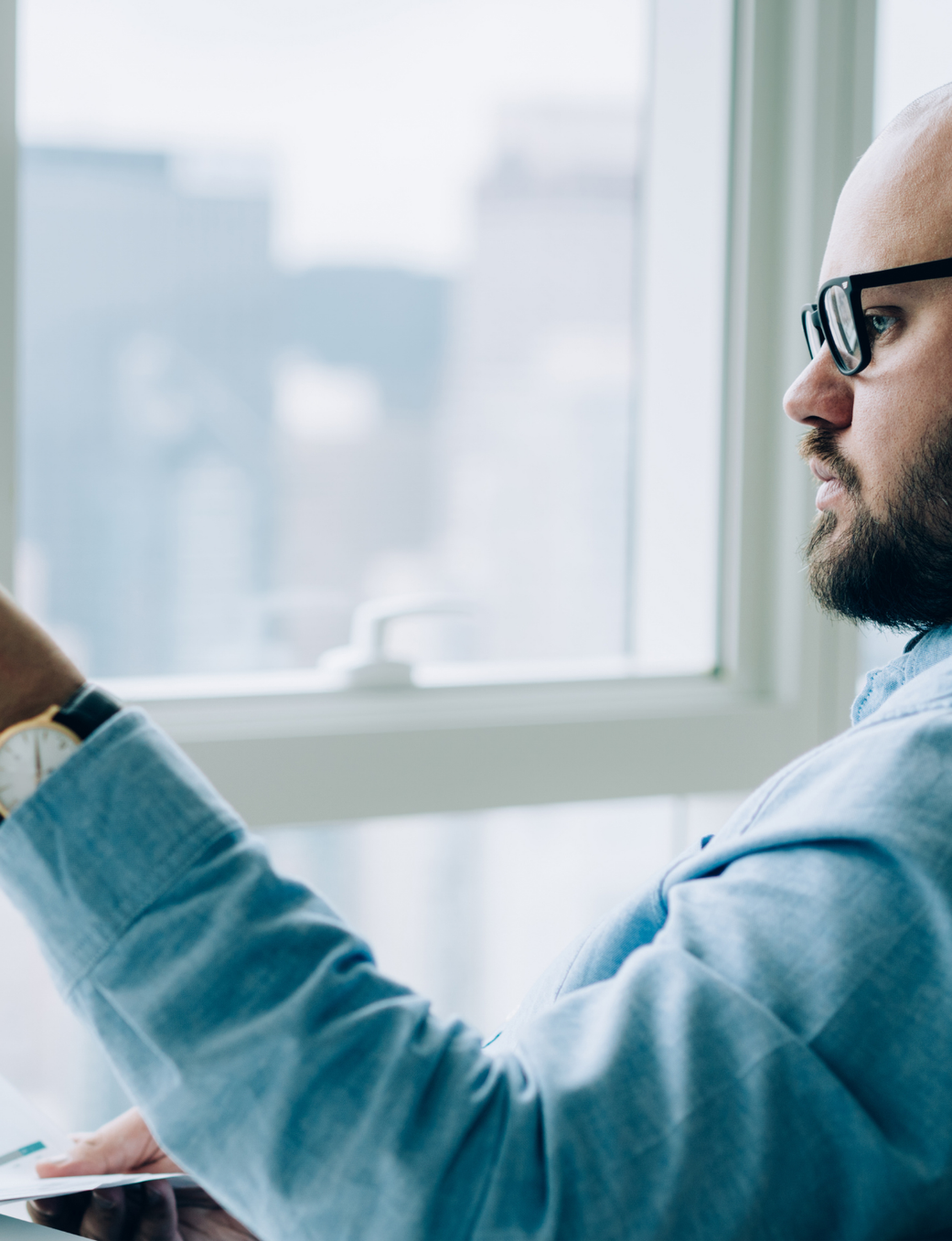 Bald man with a beard and glasses reviews documents while seated by a window.