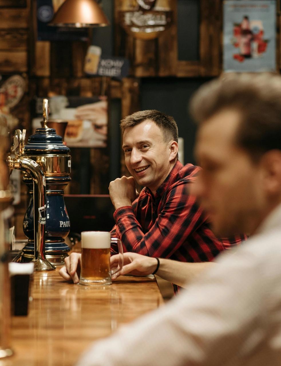 A man in a red plaid shirt smiles while holding a glass of beer at a bar.