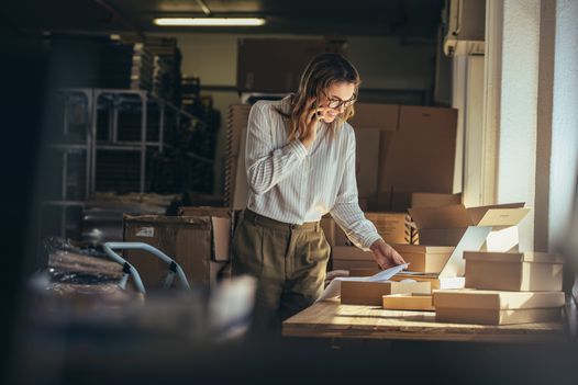 Smiling woman talking on phone while checking papers next to shipping boxes.