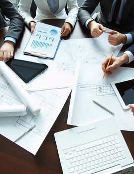 Professionals' hands review blueprints, charts, and digital devices at a meeting table.