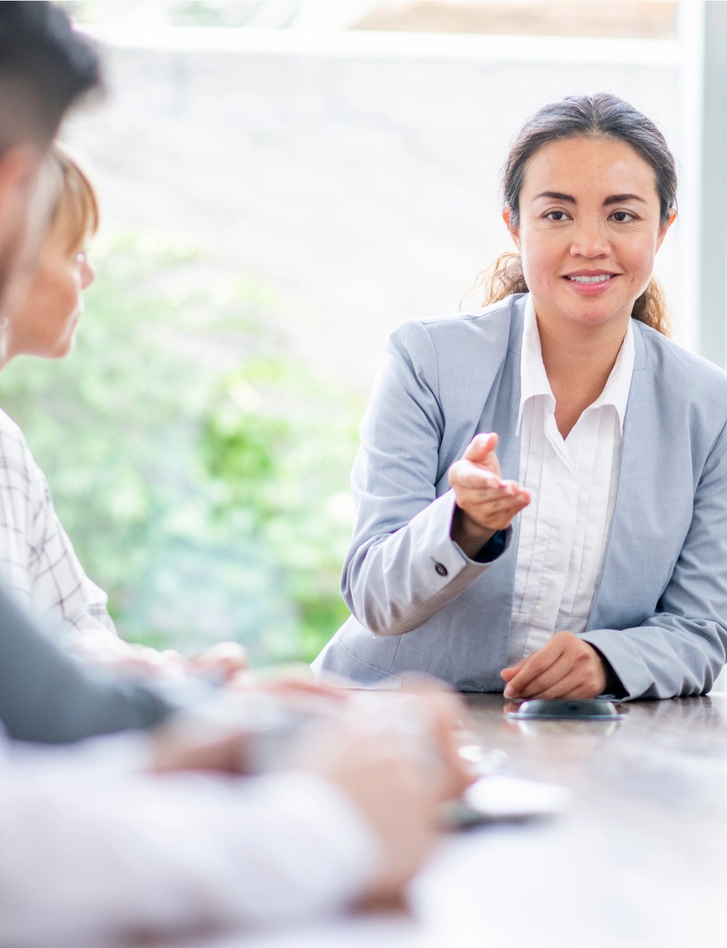 A smiling businesswoman in a suit gestures while speaking at a meeting.