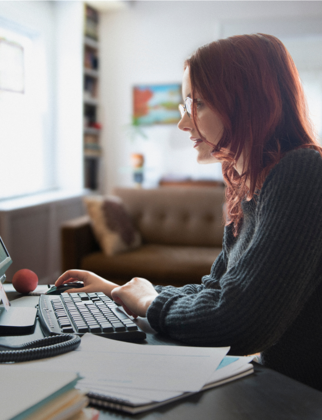 A woman with red hair and glasses works on a desktop computer at home.