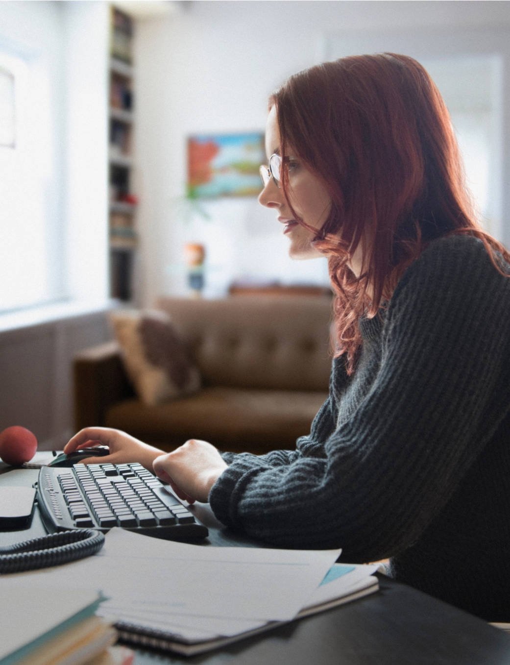 A red-haired woman with glasses sits at a desk, working on a desktop computer.