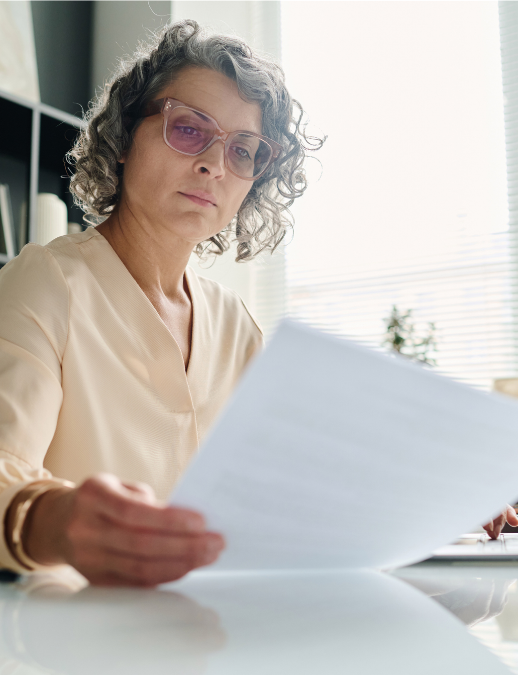 A professional woman with grey curly hair and pink glasses sits at a desk, intently reading papers with a laptop nearby.