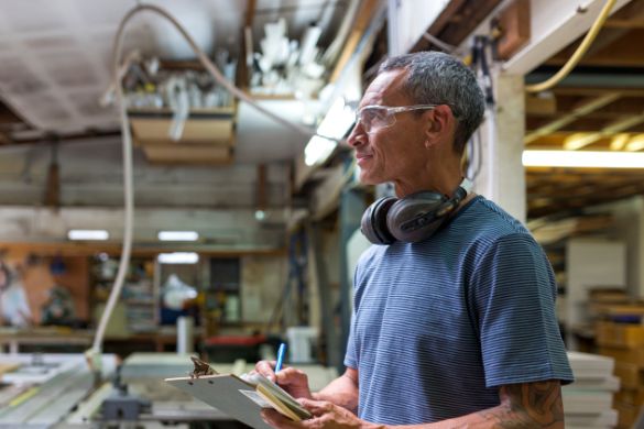 a man wearing headphones and safety glasses is holding a clipboard in a workshop .