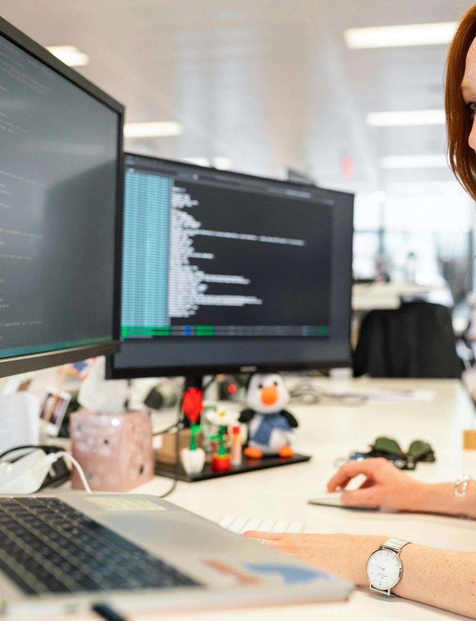 A woman with red hair works at a desk, looking at multiple computer screens displaying code.