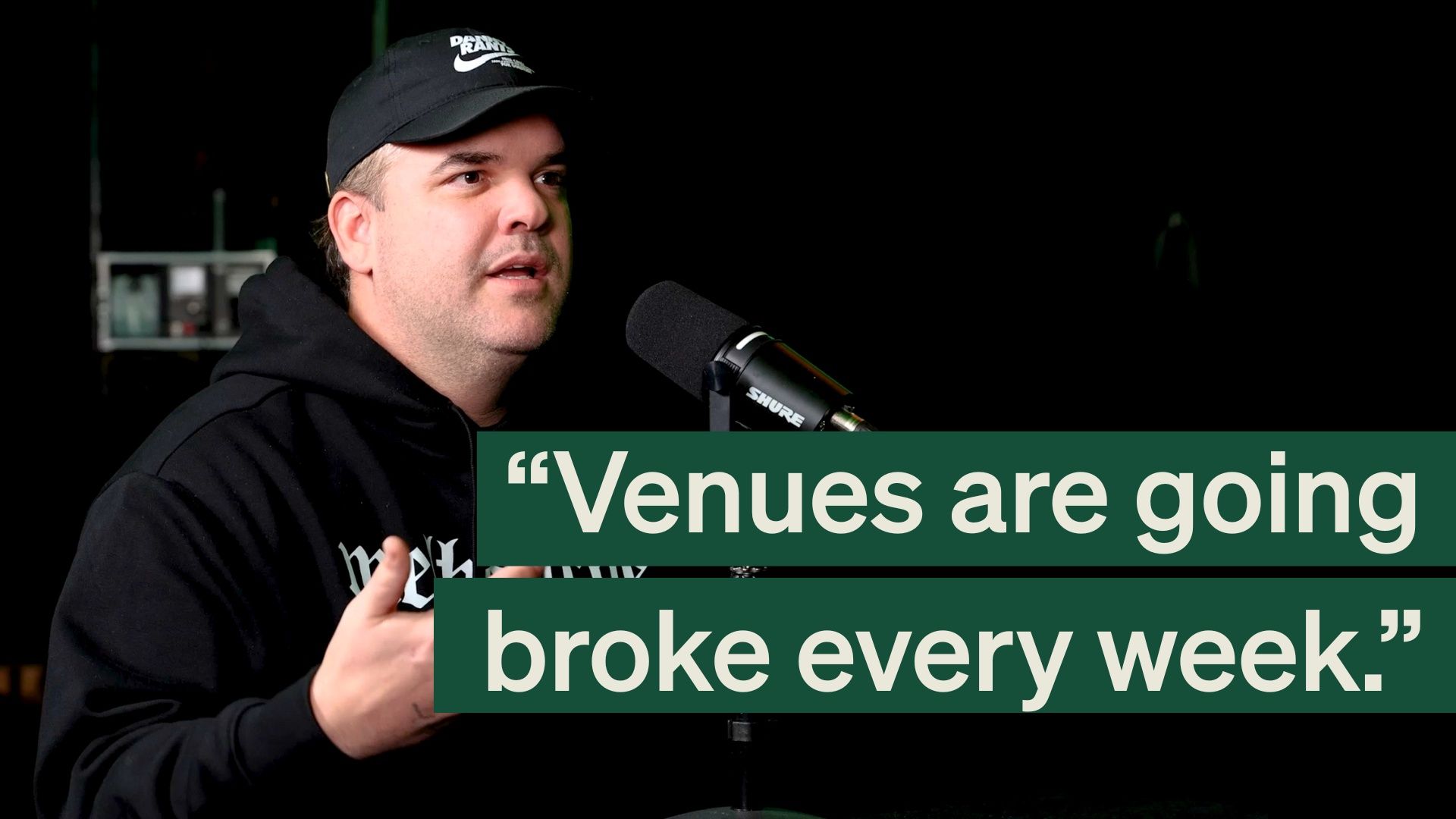 A man in a black cap and hoodie speaking into a microphone, with a text overlay reading "Venues are going broke every week."