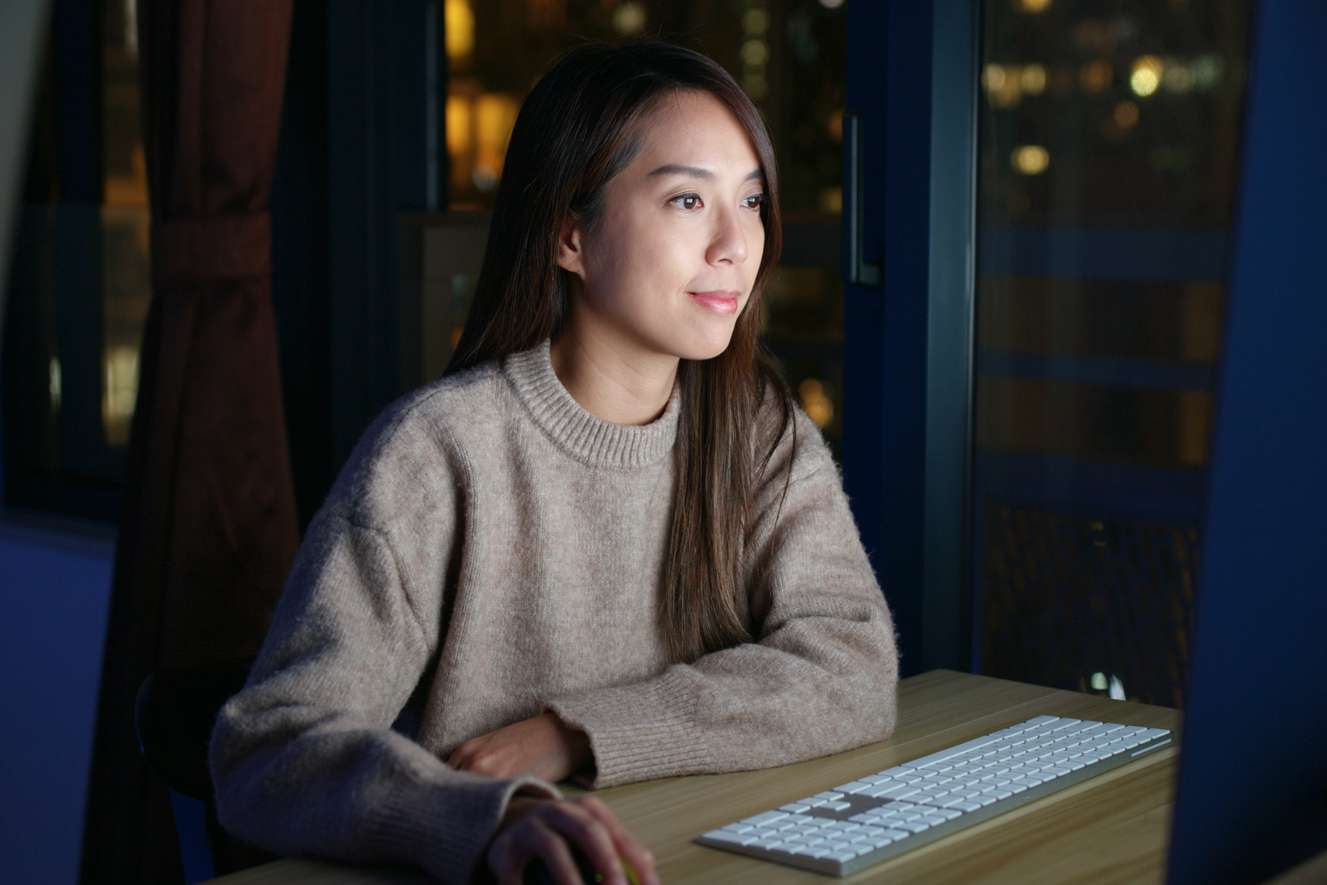A woman works at a computer at night, with city lights in the background.