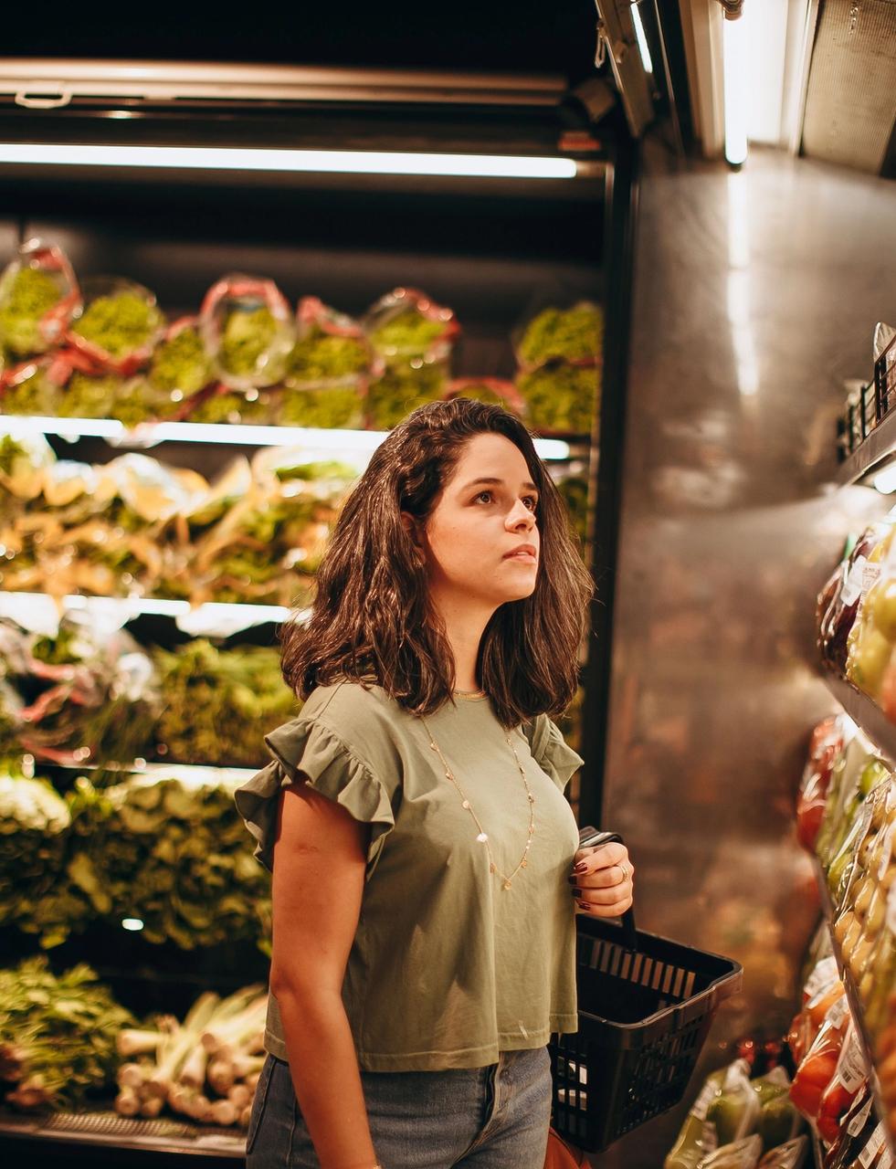 A woman with a shopping basket looks at items on a supermarket shelf.