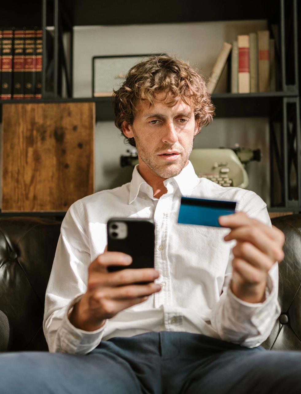 Man on a couch using his smartphone and holding a credit card.