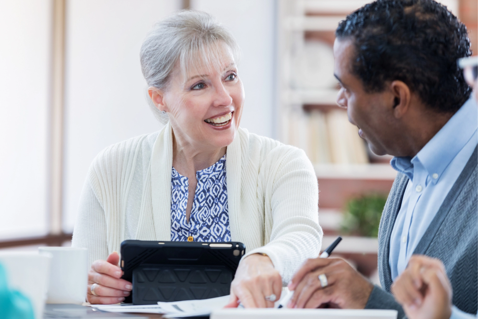 A smiling older woman with a tablet looks at an older Black man who is looking at documents.