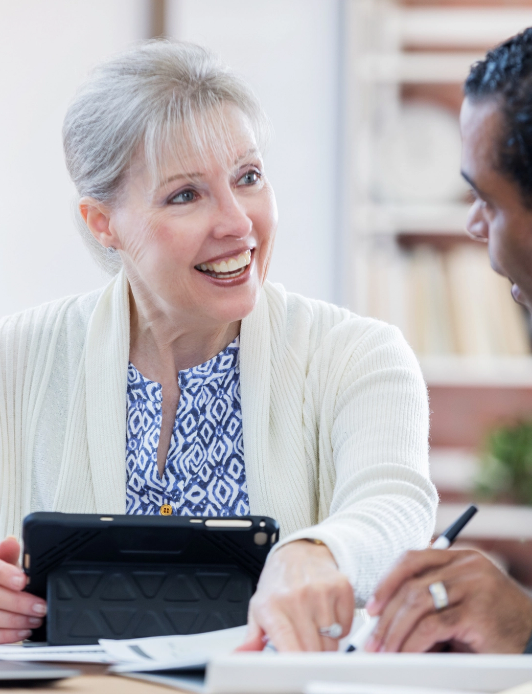 A smiling older woman with a tablet looks at an older Black man who is looking at documents.