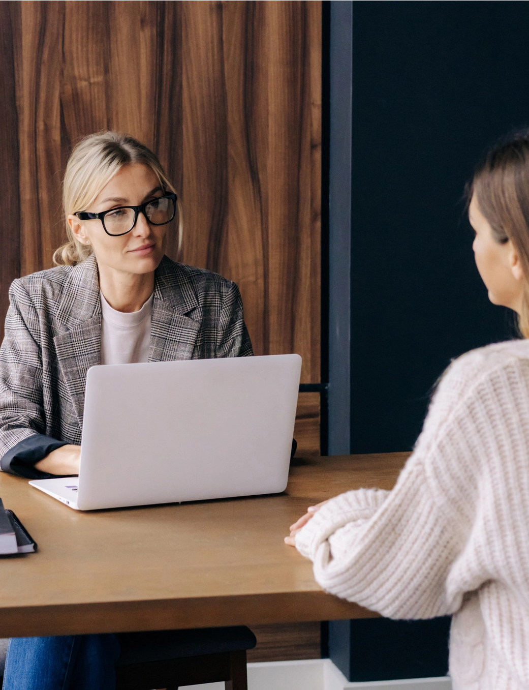 A woman with glasses at a desk with a laptop, interviewing another woman.