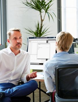 A man in a white shirt gestures while speaking to a colleague at a desk.