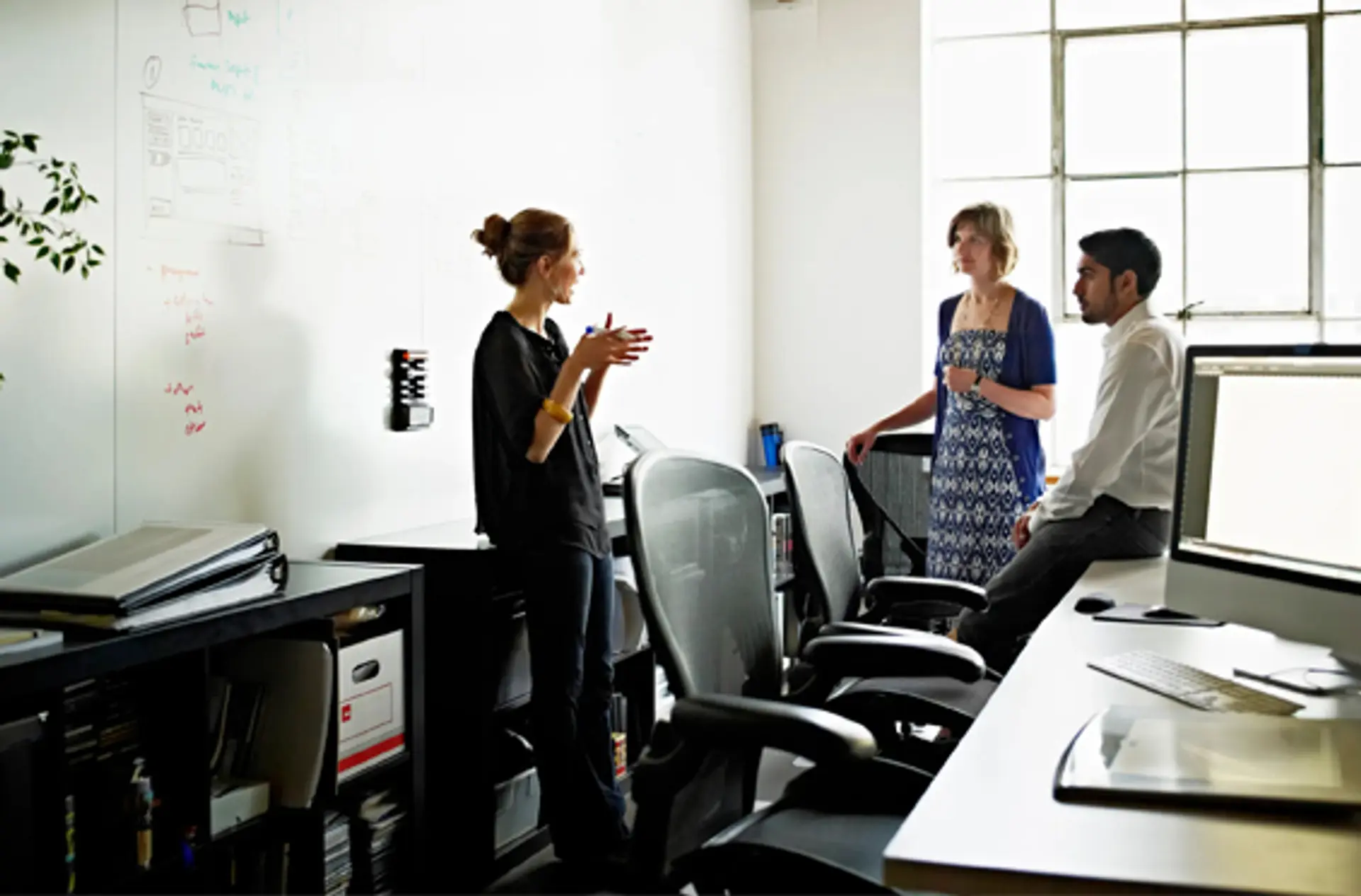 a group of people are standing in an office talking to each other .